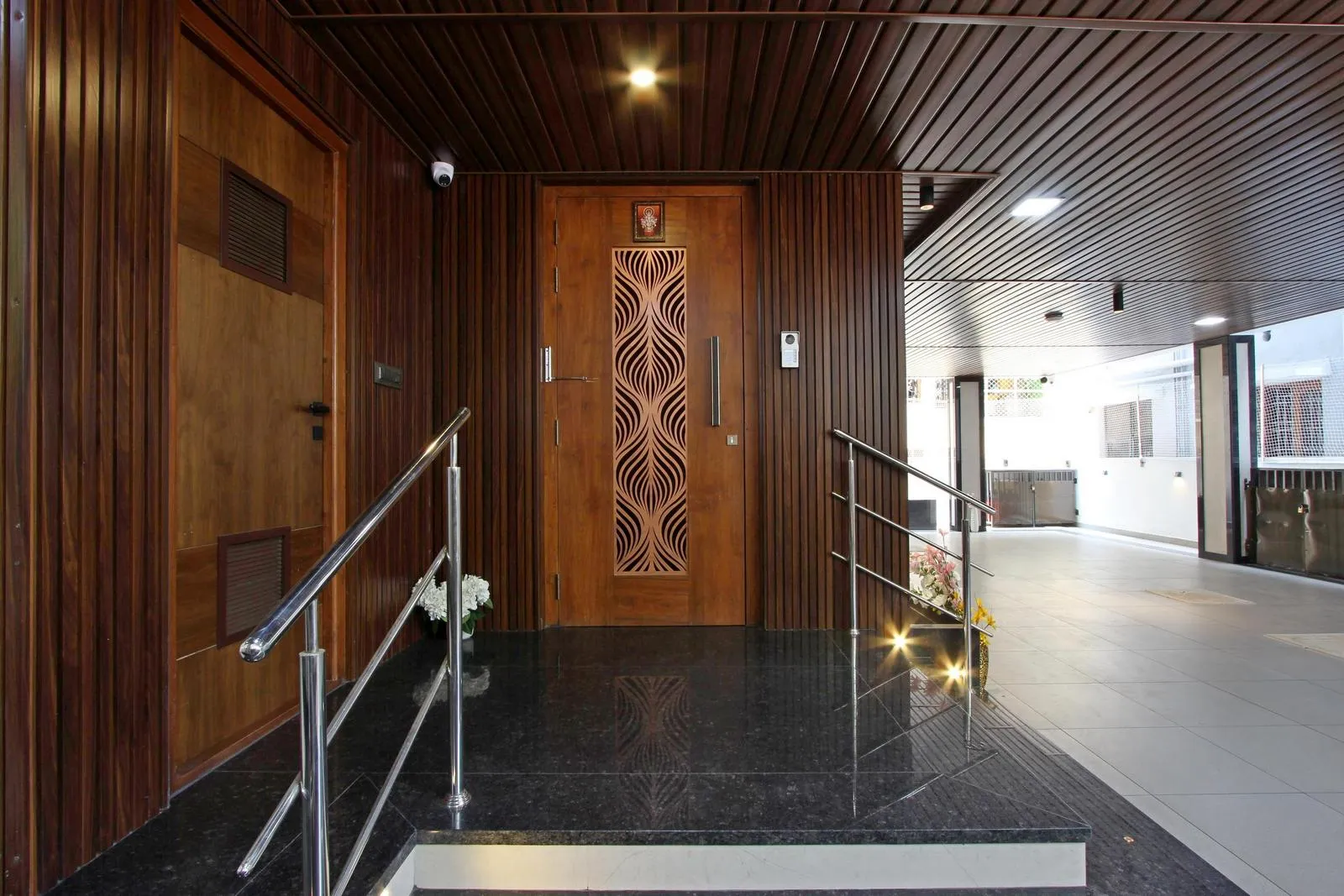 The verandah is a semi-enclosed transitional space with a central wooden door, polished granite flooring, and walnut-toned panels. Stainless-steel handrails.