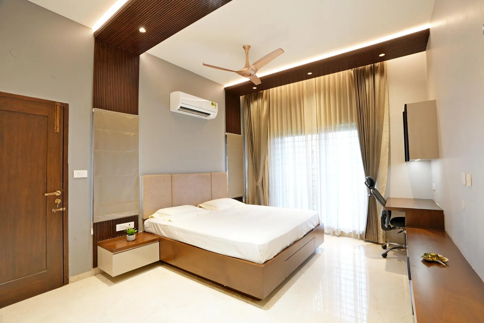 Bedroom ( 12'0" x 17'0" sq.ft ) a rectilinear layout with a central bed, balcony-access sliding door and window, and integrated study corner. Polished marble flooring, teak wood furniture, wooden ceiling panels, and soft beige curtains create a bright, functional, and elegant space.