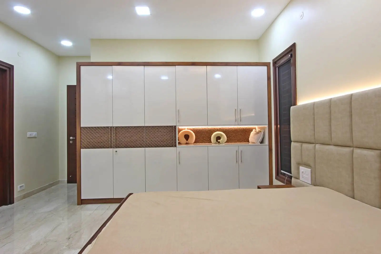 Contemporary bedroom featuring a beige upholstered bed, built-in cabinetry with brown trim, decorative lighting, and large windows, designed for comfort and functional elegance.