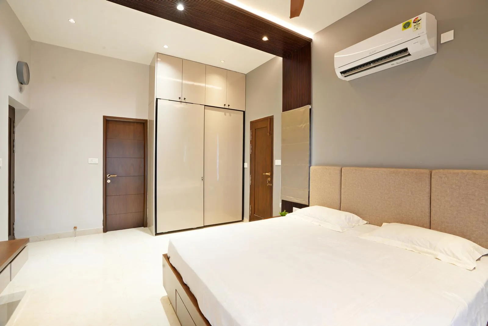 High-gloss wardrobe shutters create a bright, warm, and expansive bedroom. Layered ceiling lighting, handle-less cabinetry, and integrated wooden accents enhance functionality, visual depth, and modern minimalist elegance.