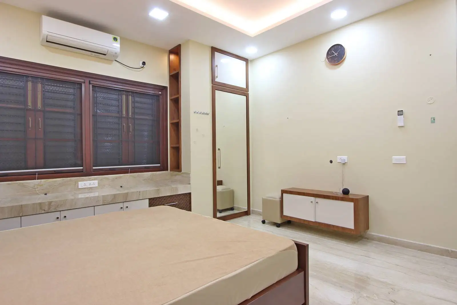 A minimalist bedroom with a beige bed, large windows with blinds, mirrored wardrobe, and wall-mounted wooden cabinet, designed for a clean, functional, and well-lit living space.