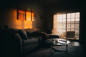 Dimly lit living room with a gray sectional sofa, a glass coffee table with books and a candle, and a sliding glass door leading to a balcony with outdoor chairs, illuminated by warm sunset light casting shadows on the wall and artwork above the sofa.