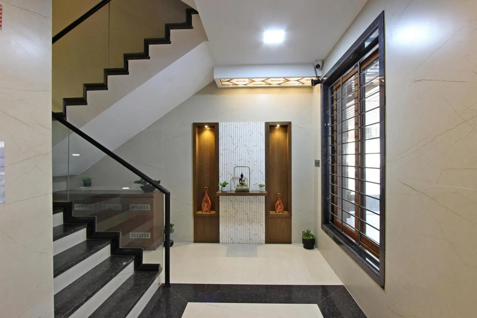 The lobby links the staircase and main circulation, with a decorative niche, large window for natural light, marble-textured walls, contrasting cream and black flooring, and warm wooden accents.