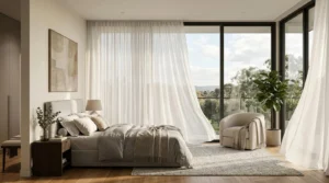 Modern bedroom with a large bed featuring neutral-colored bedding and multiple pillows, a wooden nightstand with a lamp and a glass vase with greenery, sheer curtains blowing in the breeze, a cozy armchair with a throw blanket, a large potted plant, and floor-to-ceiling windows showing a view of trees and a cloudy sky outside.