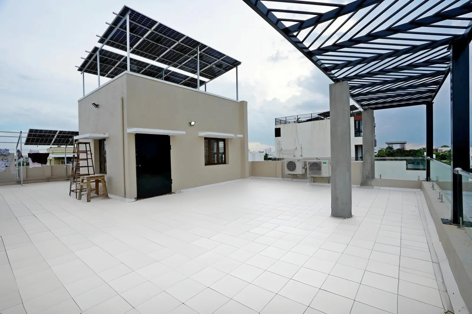 The terrace is planned as an open recreational space with a rectilinear layout, where a pergola defines a semi-shaded zone and adds architectural rhythm. Light-toned flooring, neutral walls, and a black metal pergola create a clean, modern rooftop environment that balances functionality, sustainability, and visual clarity.