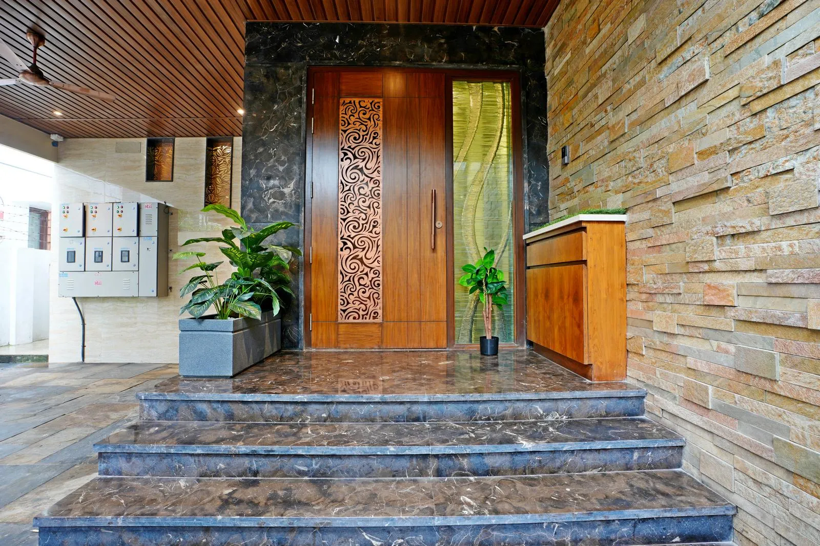 The 8'6" × 4'0" sq. ft verandah features marble-clad steps, a central wooden door with metal inlay, and side glazing for natural light. Polished marble flooring, sandstone walls, wooden ceilings, and symmetrical planters create a grand, elegant, and inviting entry space.