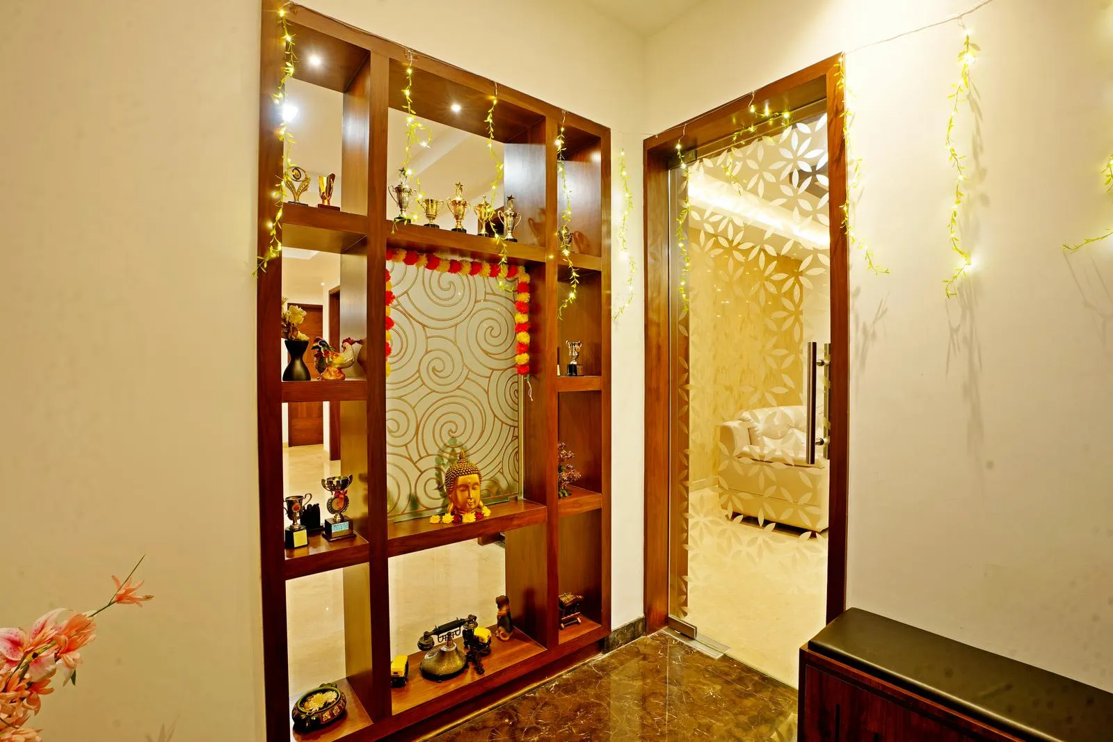 The 8'0" × 7'0" sq. ft foyer connects the entrance to the living area, featuring a custom wooden display unit and a glass partition with laser-cut patterns. Polished marble flooring, walnut shelving, and integrated lighting create a warm, elegant, and visually engaging transition space.
