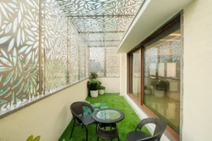Balcony with artificial green grass flooring, three black wicker chairs, and a round glass-top wicker table, featuring decorative laser-cut leaf-patterned panels on the sides and ceiling, with three potted plants in the corner and a large window reflecting the patterned panels.