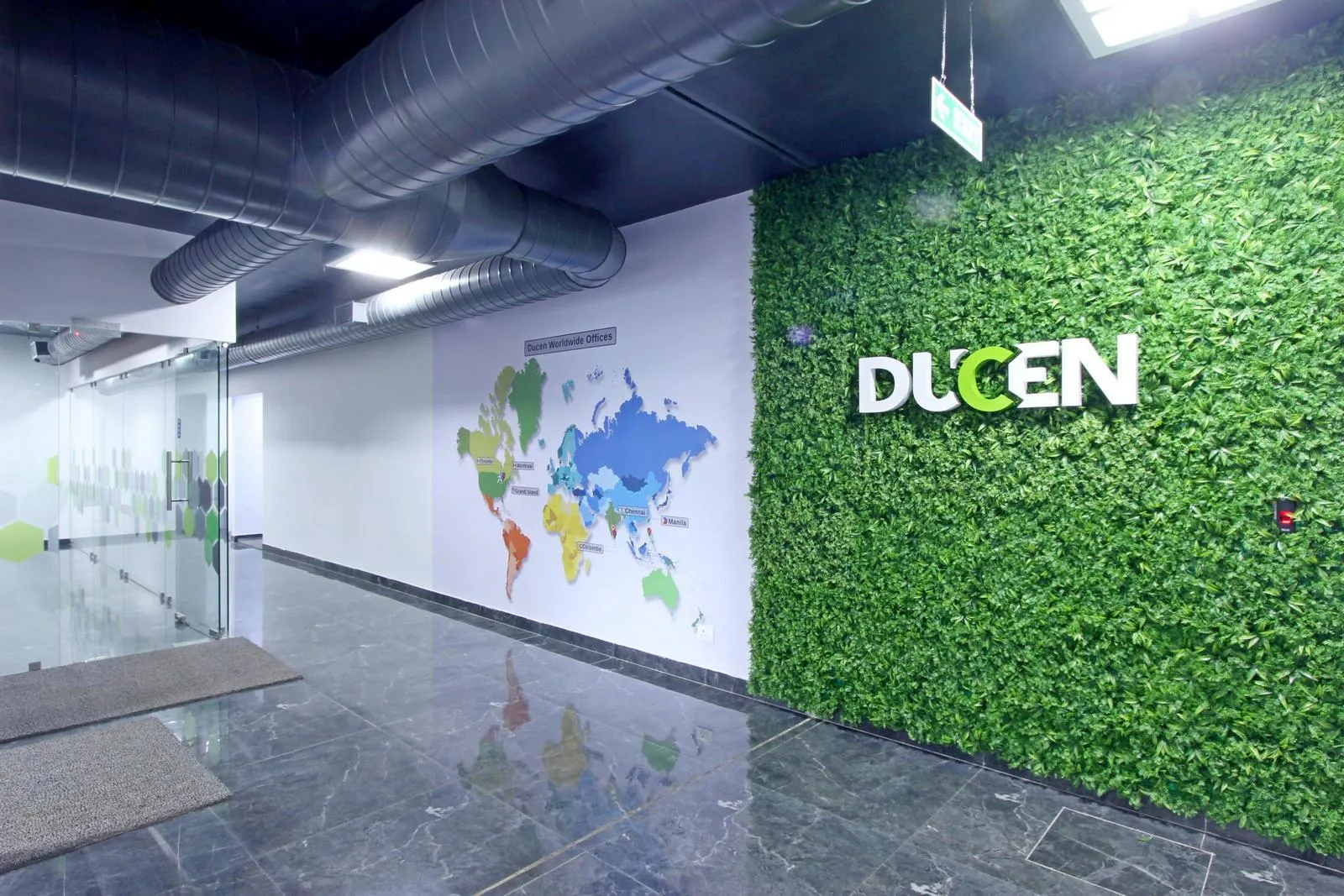 The 4th-floor reception features a vertical green wall with 3D logo signage, world map graphics, and frosted glass partitions, creating a modern, welcoming corporate entry. Dark grey marble flooring, matte black ceilings, and recessed LED lighting enhance professionalism, spatial clarity, and visual impact across the floor.