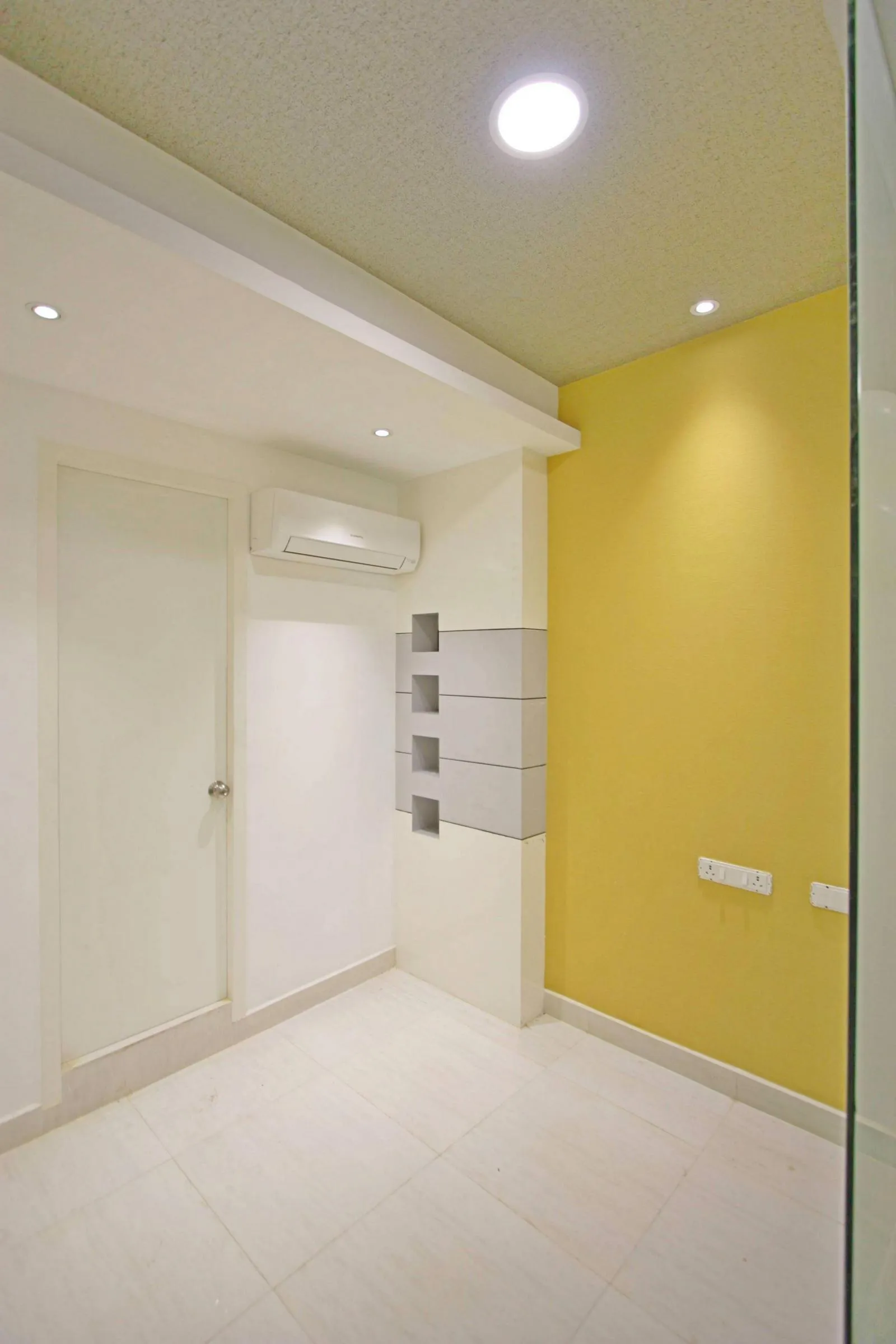 The room has a simple, rectilinear layout with neutral floors and walls, highlighted by a mustard yellow feature wall. Recessed lighting, a detailed ceiling, and built-in niches add depth, functionality, and a modern, clean look.