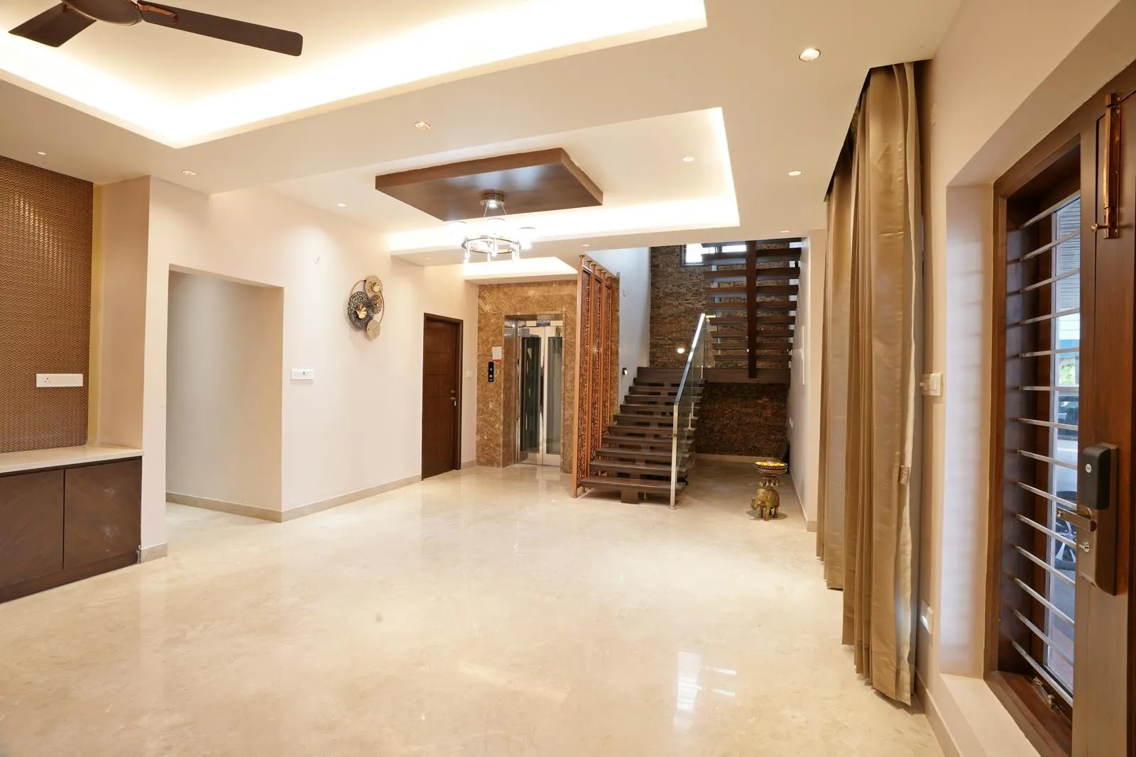 Staircase & Pebble Garden features a wooden staircase with glass railing over polished marble flooring, complemented by a pebble garden underneath that adds texture and natural charm. Layered lighting, stone-clad walls, and dark wood ceiling panels enhance depth, warmth, and visual interest.