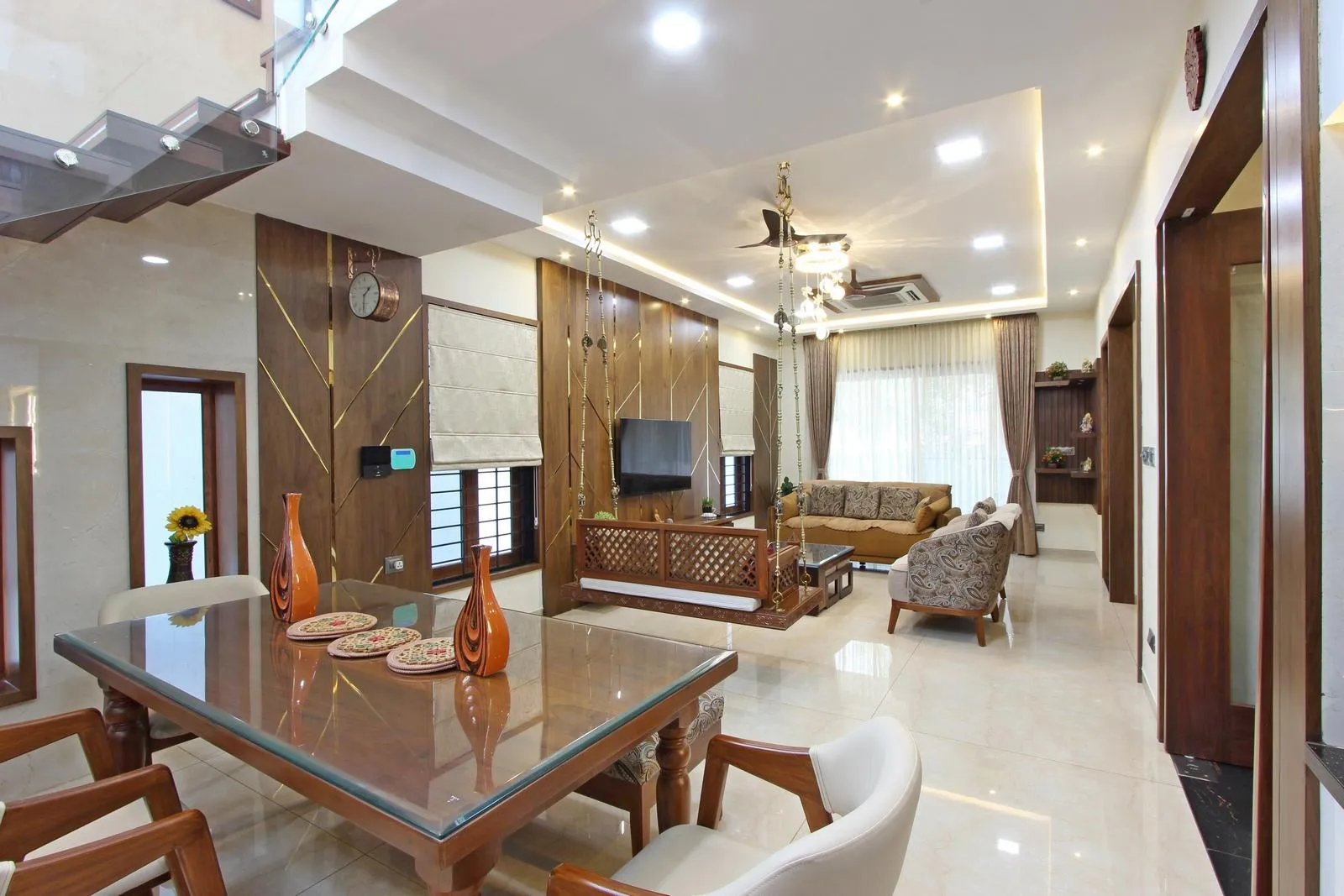 Modern living room and dining area with beige marble flooring, wooden furniture, glass-top dining table with orange vases, patterned armchairs, a matching sofa set, a ceiling fan, recessed lighting, and a swing seat suspended from the ceiling near the TV wall paneling with windows and curtains.