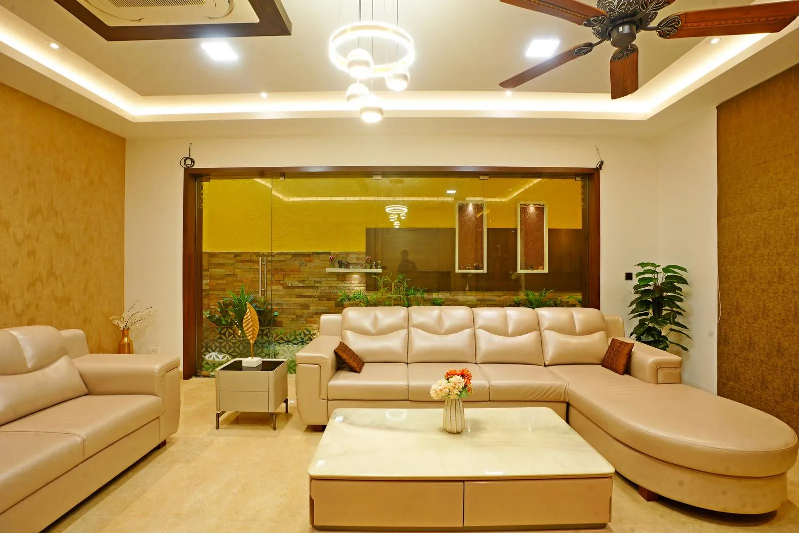Modern living room with beige leather sectional sofa, matching armchair, marble-top coffee table with flower vase, side table with decorative sculpture, ceiling fan, and large glass window showcasing indoor plants and yellow-tinted wall.