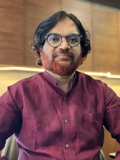 Middle-aged man with glasses, black wavy hair, and a reddish beard, wearing a maroon button-up shirt, sitting indoors with a wooden background.