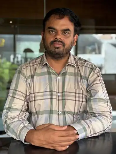 Man with short black hair and beard wearing a light-colored plaid shirt sitting at a table with hands clasped, indoor background with glass and blurred objects