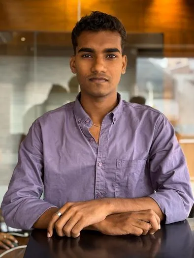 Young man with short curly hair wearing a purple button-up shirt, sitting at a table with hands folded, in an indoor setting with warm lighting and glass walls in the background.