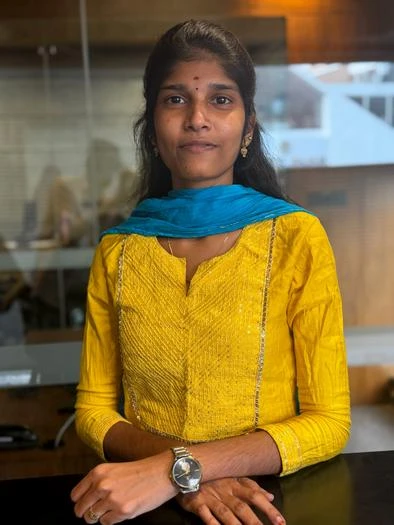 Young woman with long dark hair wearing a yellow traditional outfit and a blue scarf, standing indoors with her arms crossed and a watch on her left wrist.