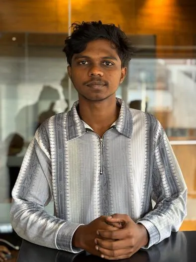 Young man with short curly hair wearing a gray patterned long-sleeve shirt, standing indoors with hands clasped in front and a calm expression