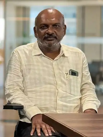 Middle-aged man with a bald head and gray beard wearing a light-colored checkered shirt sitting at a wooden table in an office environment.