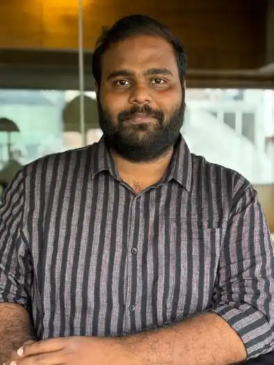 Man with a beard wearing a dark striped button-up shirt, standing indoors with a glass window and blurred background behind him.