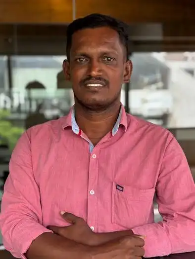 Man with short black hair wearing a pink button-up shirt, standing indoors with his arms crossed against a background of glass walls and blurred office furniture.