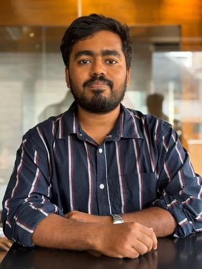 Portrait of a man with a beard and mustache wearing a navy blue striped shirt, sitting at a table with his arms resting on it against a blurred indoor background with wooden tones.