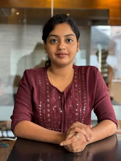 Woman with dark hair wearing a maroon embroidered top sitting at a table with hands clasped, looking at the camera, with a blurred indoor background.