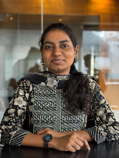 A woman with dark hair tied back, wearing a black and white patterned traditional outfit, sitting at a table with hands folded and smiling slightly against a background with glass and warm-toned panels.