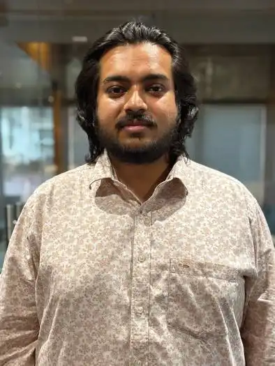 Portrait of a man with medium-length black hair and a beard, wearing a light-colored floral patterned button-up shirt, standing indoors with a blurred background.
