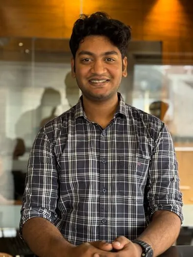 Smiling young man with dark curly hair wearing a black and white checkered shirt standing indoors with a wooden wall and window in the background.