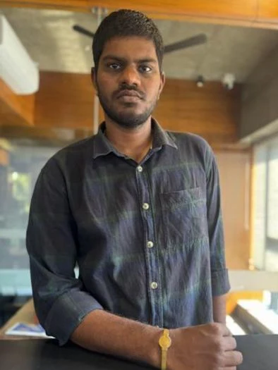 A man with short hair and a beard wearing a dark button-up shirt with rolled-up sleeves, standing indoors with wooden paneling and a ceiling fan in the background, resting his arm on a surface.