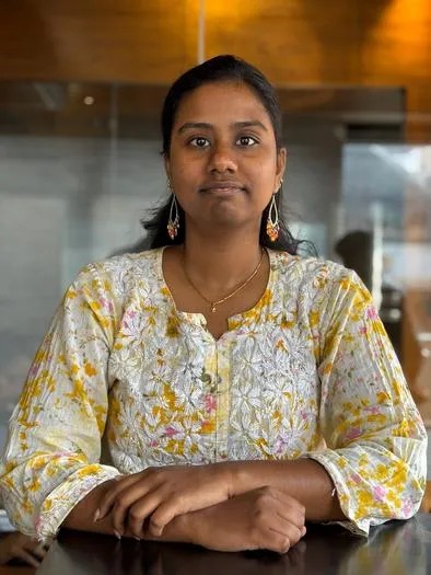 A woman with dark hair tied back, wearing a white and yellow floral patterned top, sitting at a table with hands folded, against a background with warm wooden tones and glass reflections.