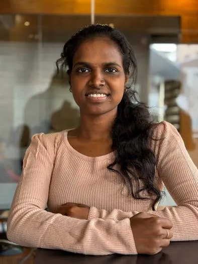 Smiling woman with long dark hair wearing a light pink ribbed long-sleeve shirt sitting at a table with folded arms in a warmly lit indoor setting.