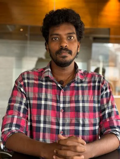 Man with curly hair and beard wearing a red and blue checkered shirt, sitting indoors with hands clasped on a table, looking directly at the camera.