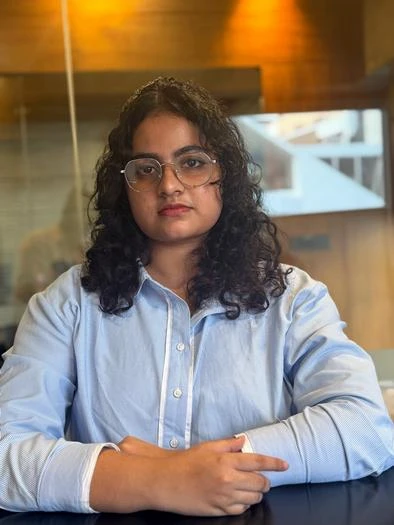 Young woman with curly black hair and glasses wearing a light blue button-up shirt, sitting at a table with her hands folded, indoors with warm lighting and a blurred background.