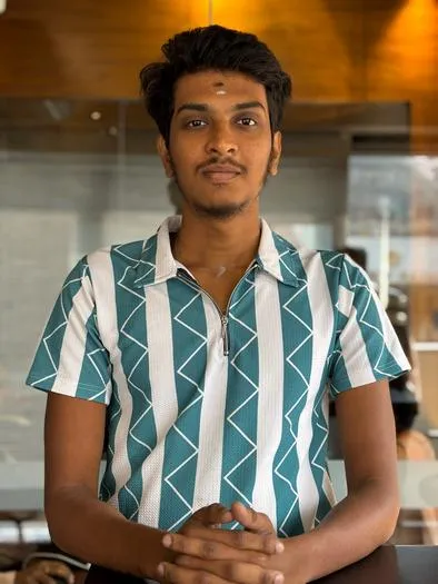 Young man with medium skin tone and short black hair wearing a teal and white vertical striped shirt with a geometric pattern, standing indoors with folded hands and looking at the camera.