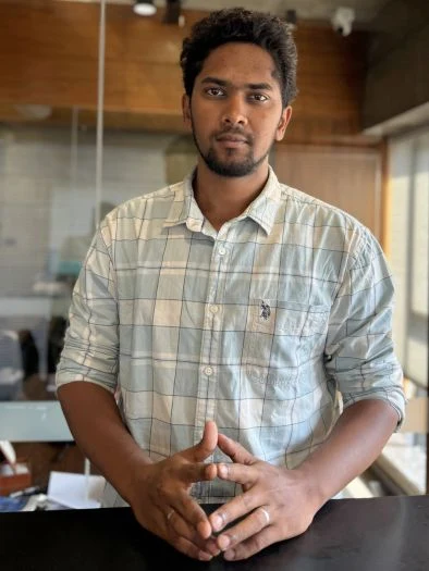 Young man with dark hair and beard wearing a light checkered shirt standing indoors with hands clasped on a table in front of him, in a room with wood and glass background.