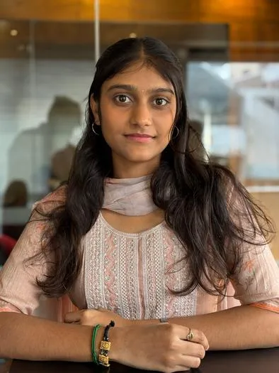 Young woman with long dark hair wearing a light pink embroidered traditional outfit, sitting with her hands folded on a table indoors in front of a blurred background.