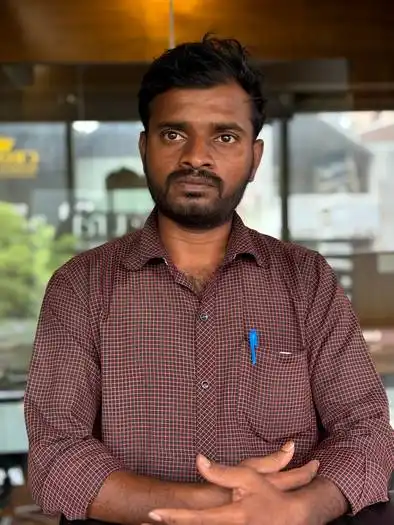 Man with dark hair and beard wearing a checkered maroon shirt with rolled-up sleeves, standing indoors with a neutral expression and a pen in his shirt pocket.