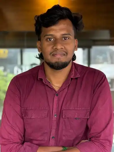 Portrait of a man with short black hair and a beard, wearing a maroon button-up shirt, standing with arms crossed against a blurred indoor background.