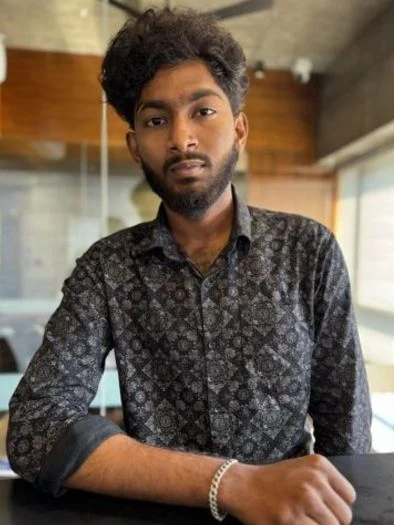 Young man with curly hair and a beard, wearing a patterned dark shirt with rolled-up sleeves, sitting at a table indoors with a casual expression