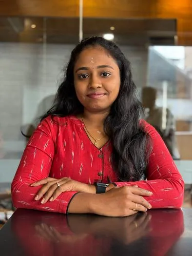 Young woman with long dark hair wearing a red patterned top sitting at a table with arms crossed, smiling softly, with a blurred indoor background.