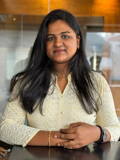 Young woman with long black hair wearing a cream-colored embroidered top, sitting at a table with hands clasped, in an indoor setting with warm lighting and blurred background.