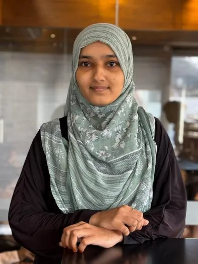 Portrait of a young woman wearing a light green patterned hijab and a black long-sleeve top sitting with hands folded on a table, with a blurred indoor background.