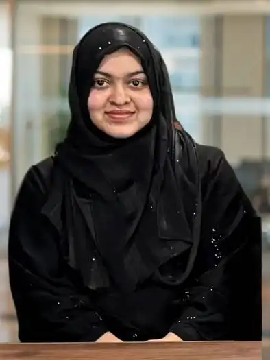 Portrait of a woman wearing a black hijab and black outfit, smiling gently, seated indoors with a blurred background.