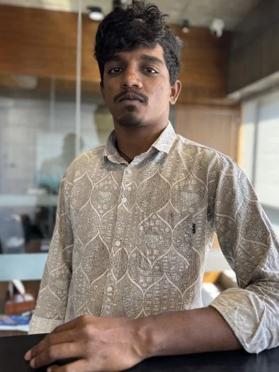 Young man with curly black hair wearing a patterned light gray button-up shirt, standing indoors near a window with a serious expression.