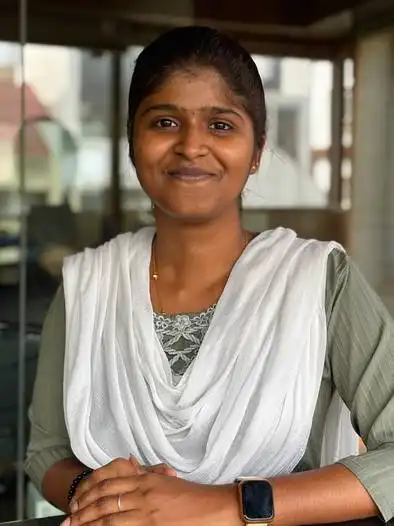 Portrait of a smiling woman with dark hair tied back, wearing a green top and a white scarf draped over her shoulders, standing indoors with blurred background.
