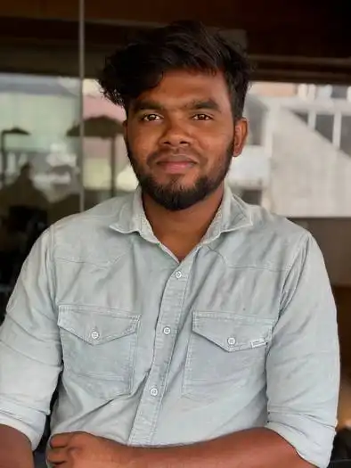 Young man with short curly hair and beard wearing a light grey button-up shirt, standing indoors with a glass wall and building in the background.
