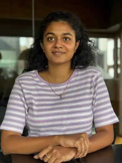 Woman with curly black hair sitting at a table, wearing a purple and white striped short-sleeve top and a necklace, in an indoor setting with blurred background windows.