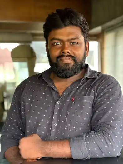 Man with dark hair and beard wearing a dark gray button-up shirt with small patterns, standing indoors with arms resting on a surface and a window background.